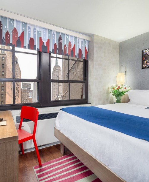 king guest bed with white linens, navy draped end blanket, red chair at desk, and view of empire state building